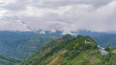 yemyeşil dağlar bulutlu bir gökyüzüne doğru yükselirler, vadilerde süzülen sis ve yamaçta tünemiş küçük bir tepe kompleksi.