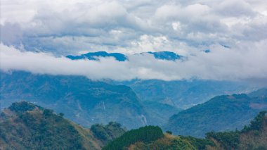 Bulutlarla kaplı dağ sıraları, bulutlu bir gökyüzünün altında mavi ve yeşil tepeler..