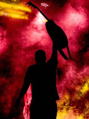 Grubun solisti, girdap gibi kırmızı ve sarı dumanlarla çevrili bir konser sırasında gitarını kaldırır. Silueti, etrafındaki renkli sisi aydınlatan ışığın çarpıcı ve enerjik bir şekilde yansıttığı canlı arkaplan karşısında karanlık görünüyor. 