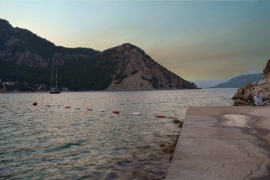 Alacakaranlık yaklaşırken arka planda bir körfez, kayalar ve Turunc (Türkiye) tatil köyü manzarası.