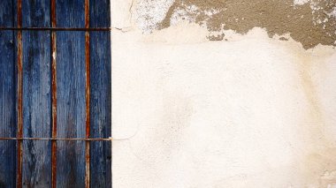 Parmaklıklı pencereli beyaz duvar ve ön cephesinde mavi ahşap panjurlar.