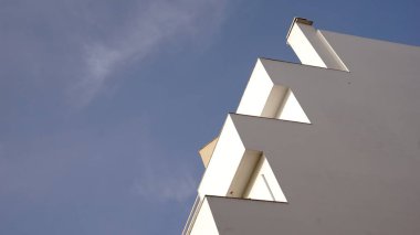 Açık mavi gökyüzüne karşı keskin açısal şekillere sahip beyaz bir binanın minimalist görüntüsü.