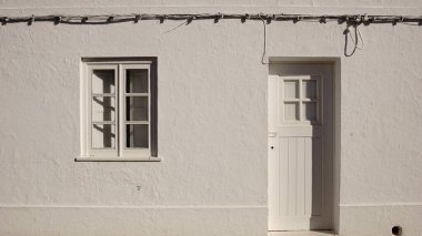 Küçük pencereli ve ahşap kapılı, görünen elektrik kablolarından oluşan basit bir beyaz duvar..