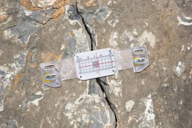 Deep crack in a natural old stone wall cause due structural subsidence of the subsoil with plastic mechanical crack meter designed to measure movement across surface cracks and joints clipart