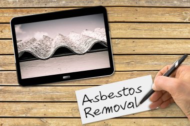 Hand writing Asbestos Removal text with a pencil on a white sheet above a wooden desk with a digital tablet showing a dangerous asbestos roof - 3D render concept image clipart