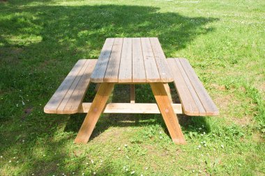 Kamu parkında banklı ahşap piknik masası yapraklı ağaçların altında gölgeli noktalar sunuyor..