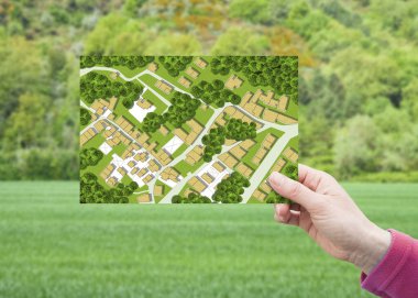 Arazi yönetimi - yeşil yerleşim alanındaki boş bir arazide, hayali bir kadastro haritasıyla ilgili bir kartpostalı elinde tutarak inşaat için kullanılabilir arazi kavramı