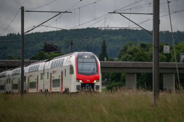 İsviçre Federal Demiryolları SBB 'nin Stadler KISS' i olarak tanımlanabilen modern, beyaz ve kırmızı çift katlı bir tren, çerçeve boyunca soldan sağa hareket ederken belirgin bir şekilde ön plana çıkıyor. Tren geliyor.