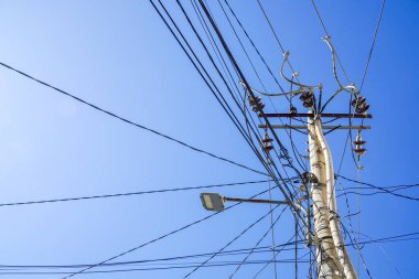 Elektrik direği ve açık mavi gökyüzü olan transformatör. Kutuplardan büyük miktarda elektrik kablosu sarkıyor..