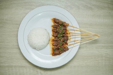 Beyaz bir tabakta fıstık soslu satay ve ahşap desenli bir masada pirinç.