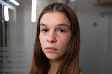 Ciddi bakışlı genç bir kızın portresi. Cilt bakımı konsepti, evinin banyosunda ve banyo ışıkları bardağa yansıyor..