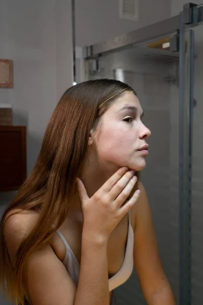 Young girl looking in the mirror looking for skin damage, pimples, irritation. she is in the bathroom of her house. skin care concept.