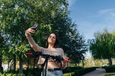 Elektrikli scooter ve mobil cihaz. Akıllı telefon kullanan genç bir kız. Şehir parkında elektrikli scooterla selfie çekiyordum. manzara fotoğrafı.