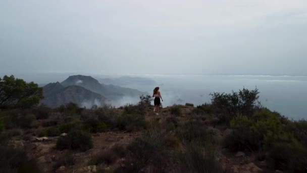 女孩从山上跑下 走到山顶 张开胳膊 空中的景色以群山和大海之间的云彩景观结束 — 图库视频影像