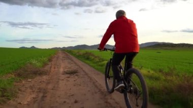 Elektrikli dağ bisikletli yaşlı bir adam yeşil bir alanda spor yapıyor. O bir yol üzerindedir ve arka planda bazı dağlar vardır. Gün batımında bulutlu bir gün. Yaşlıları aktif, hayattan zevk alan konsept.