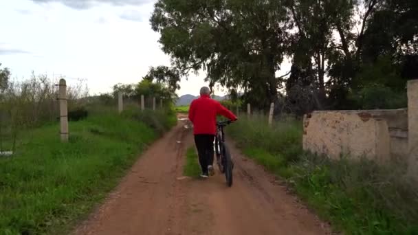 雨の日の後 老人は田舎を自転車で歩く 彼は両側に植物や木がある道路にいます 曇った日だ 積極的にコンセプトの高齢者 人生を楽しむ — ストック動画