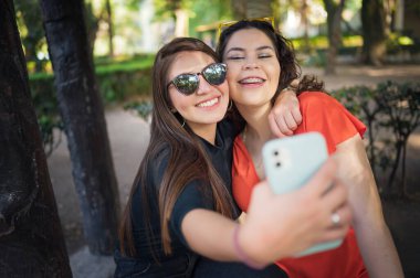 Two young latin female friends walking at the woods. Embrace each other and laughing. Friends making selfie. Two beautiful latin young women making selfie