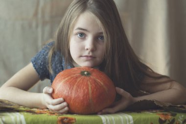 Uzun açık kahverengi saçlı ve mavi gözlü bir kız önünde parlak turuncu bir balkabağı tutuyor, düşünceli bir şekilde kameraya bakıyor. Sıcak, sonbahar temalı, yumuşak doğal ışıkla dolu sakin bir hayat..