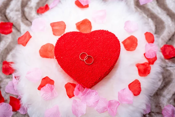 red heart-shaped box with wedding rings on top on a white pillow and rose petals around it