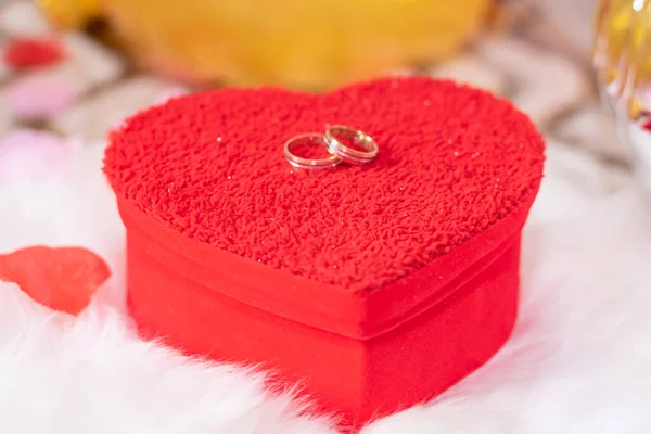 red heart-shaped box with wedding rings on top on a white pillow and rose petals around it