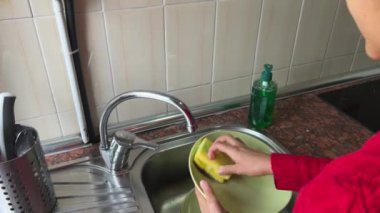 Bir insan mutfak lavabosunda bulaşık yıkıyor. Lavabo kirli bulaşıklarla dolu ve kişi onları temizlemek için sünger kullanıyor.