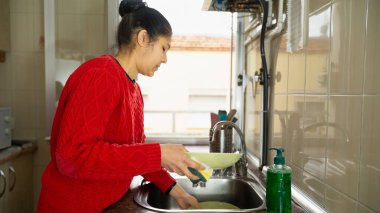 Bir kadın mutfakta bulaşıkları yıkıyor. O kırmızı bir kazak giyiyor ve yeşil bir bulaşık havlusu tutuyor.