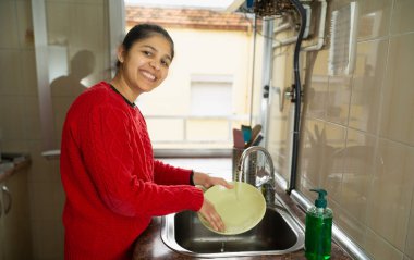 Bir kadın mutfak lavabosunda tabak yıkıyor. Gülümsüyor ve görevinden zevk alıyor.