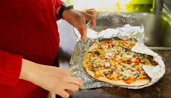 A person is holding a pizza in a tin foil. The pizza is covered in vegetables and cheese