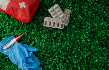 Yeşil çimenlikte kırmızı ve beyaz bir ilk yardım çantası var. Çantanın yanında bir termometre var.