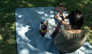Bir kadın parkta bir battaniyenin üzerinde oturuyor, kitap okuyor. Yanında bir şişe su ve bir şişe meyve suyu var.