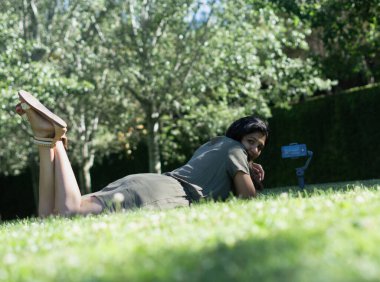 Bir kadın elinde kamerayla çimlerin üzerinde uzanıyor. Gülümsüyor ve eğleniyor.