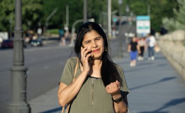 Bir kadın kaldırımda yürürken cep telefonuyla konuşuyor. Yeşil bir gömlek giyiyor ve eli kalçasında.