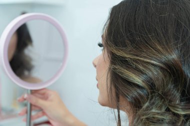 Uzun kahverengi saçlı bir kadın aynaya bakıyor. Elinde bir saç fırçası tutuyor.