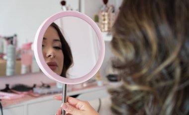 Bir kadın pembe aynaya bakıyor. O, yüzüne bir ayna tutuyor. Ayna pembe ve gümüş çerçeveli. Kadın pembe bir gömlek giyiyor.