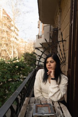 Elinde sigarayla balkonda oturan bir kadın var. Beyaz bir kazak ve saat giyiyor.