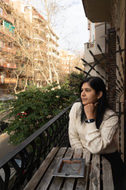 Bir kadın balkonda oturmuş şehri seyrediyor. Beyaz bir kazak ve siyah bir bileklik giyiyor.