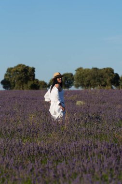 Bir kadın lavanta tarlasında yürüyor. Beyaz bir elbise ve hasır bir şapka giyiyor.
