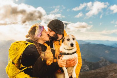 Köpekleri ile dağ yolu yaparken yağmurda öpüşen iki yürüyüşçü. Dağcı, arka planda dağ manzarası olan köpeğini kollarında tutuyor. Açık hava sporu