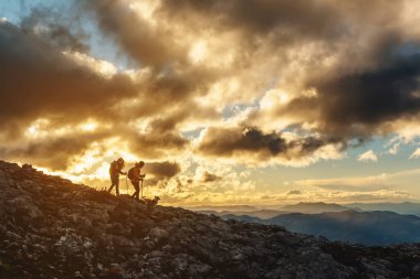 Köpekleri siluetli iki yürüyüşçü bulutlu bir günde gün batımında bir dağa iniyorlar. Bir çift yürüyüş yapıyor. spor ve açık hava macerası.