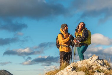 Sırt çantaları ve yürüyüş direkleriyle bir dağ zirvesine ulaştıktan sonra manzarayı seyreden iki yürüyüşçü. Erkek ve kadın yürüyüş. Açık hava sporu.