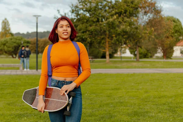 若いラテン系の女の子がスケートボードとバックパックを持って大学のキャンパスの外を歩く 学校や研究や若者文化に戻り — ストック写真