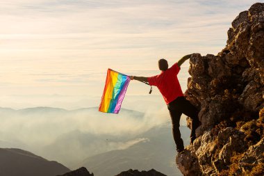 Dağcı adam bir dağda zirve yapıyor ve gün batımında gökkuşağı lgbt gurur bayrağı sallıyor. Igbt hareketi ve hak kavramı. Igbt halkının fetih hakları kavramı.