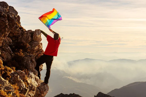 登山家の男が山で頂上を作って夕日に虹色のLgbtの誇りフラグを振っている Lgbtの運動と権利の概念です ゲイの人は山に登る スポーツと冒険 — ストック写真