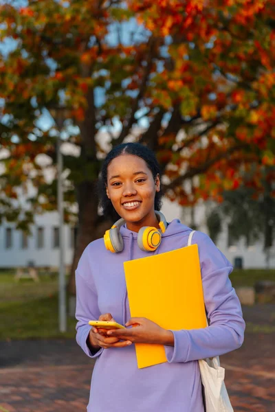 portrait of latina teenage girl, happy, holding a yellow folder, with headphones and smart phone at the school exit. Teenager with unaltered skin. student.