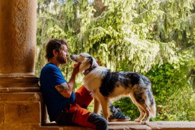 Arka planda orman olan eski bir binada, sınır köpeğinin yanında oturan genç beyaz adam. Adam evcil hayvanıyla antrenman yapıyor ve oynuyor.