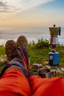 Bir dağ yürüyüşçüsünün bacaklarını gören ilk kişi güneş doğarken elinde kamp sobasıyla bir İtalyan kahve makinesinde kahve hazırlıyor. Spor ve macera. sırt çantası taşıyıcısı.