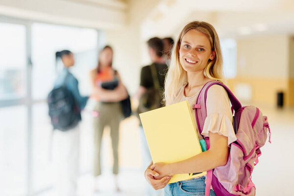 young caucasian woman, college student, posing in the campus hallway with academic materials with his classmates in the background