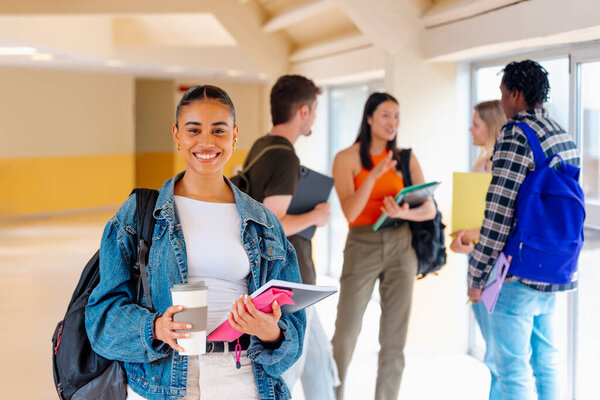 Portrait of Latina student girl looking at camera with a cup of coffee and school supplies in the hallways of the university campus with a multiracial group of classmates in the background.