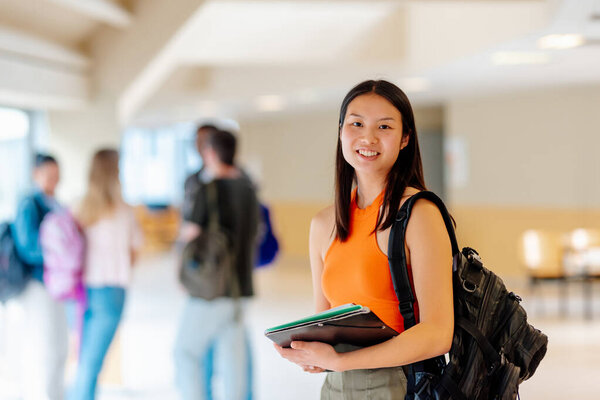 Портрет азиатской девушки с рюкзаком и школьными принадлежностями в коридорах университетского городка с одноклассниками на заднем плане.