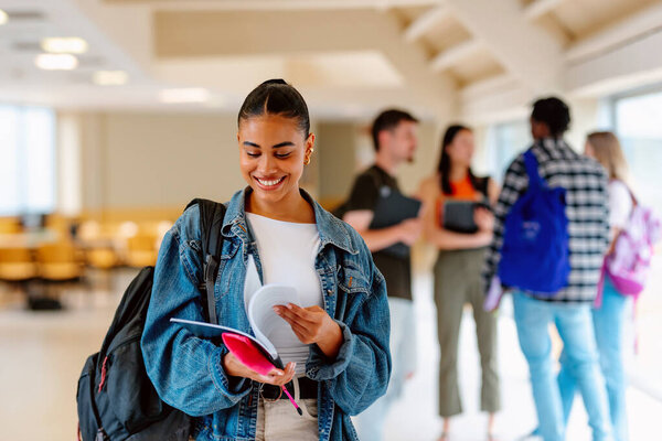 Portrait of smiling Latin girl looking at her college notes in a hallway on campus or high school with her classmates in the background. Exam time.